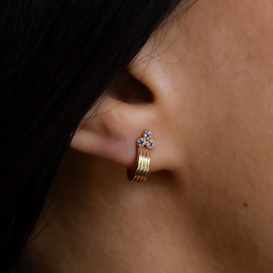 A close-up image of a person's ear wearing a gold hoop earring with three blue Iolite stones set in the hoop.