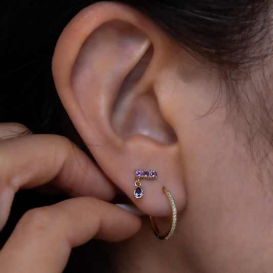 A person's hand holding a yellow gold hoop earring with a purple amethyst and a dangling pearl embellishment.