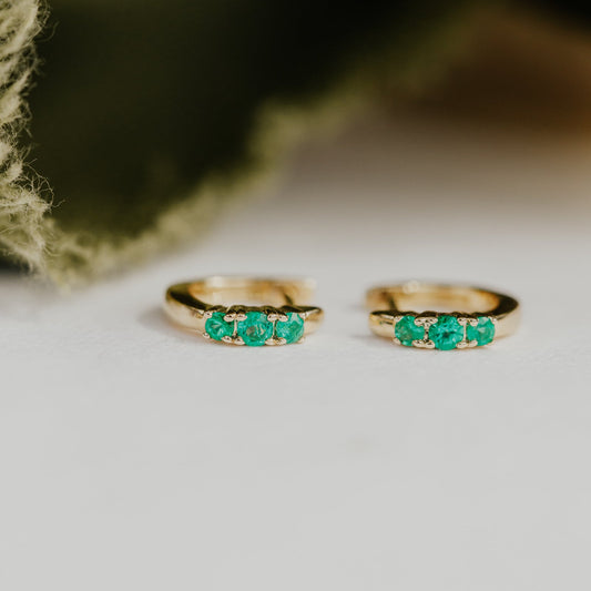 Two gold earrings with green emerald gemstones on a textured surface.