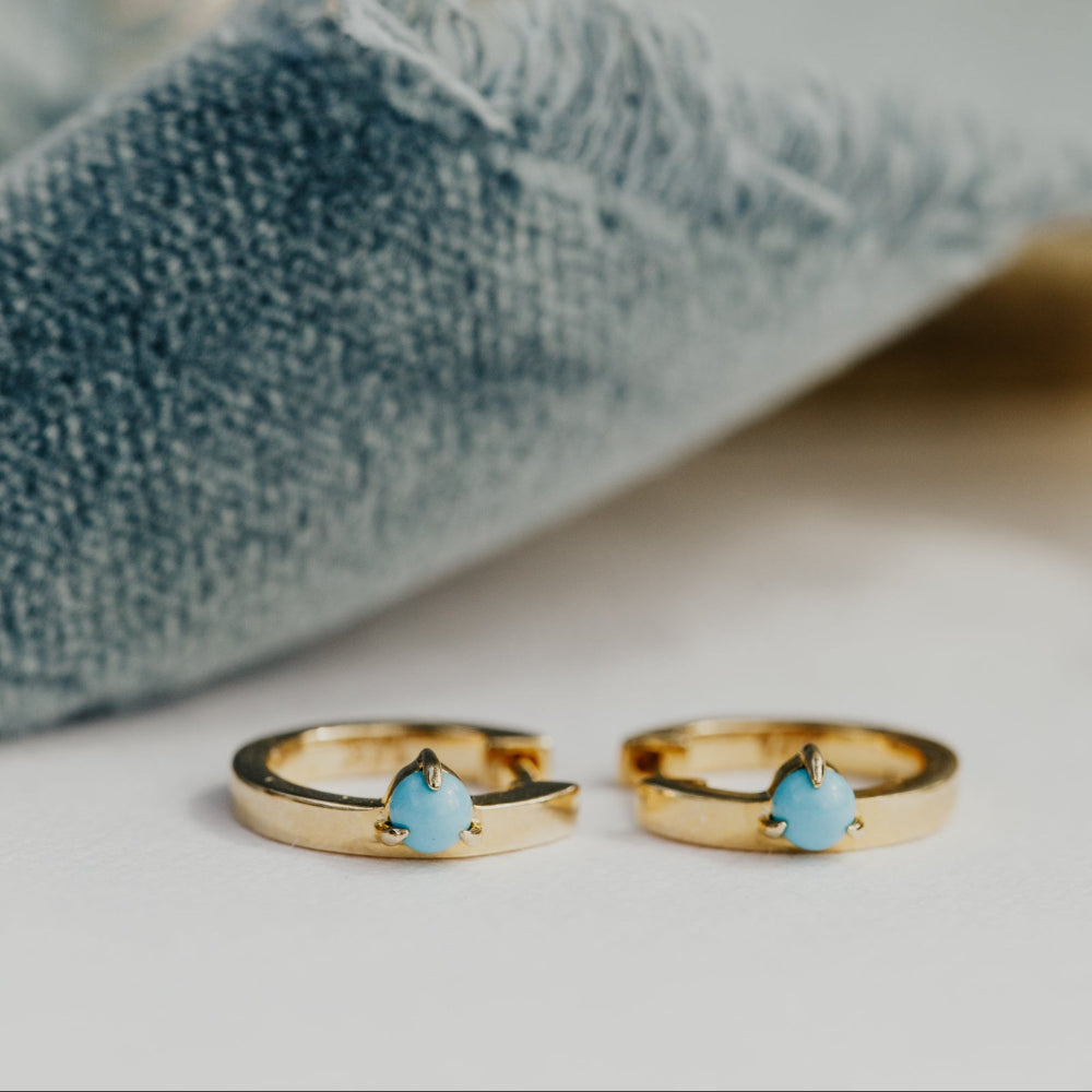 Two gold earrings with turquoise stones on a textured surface.