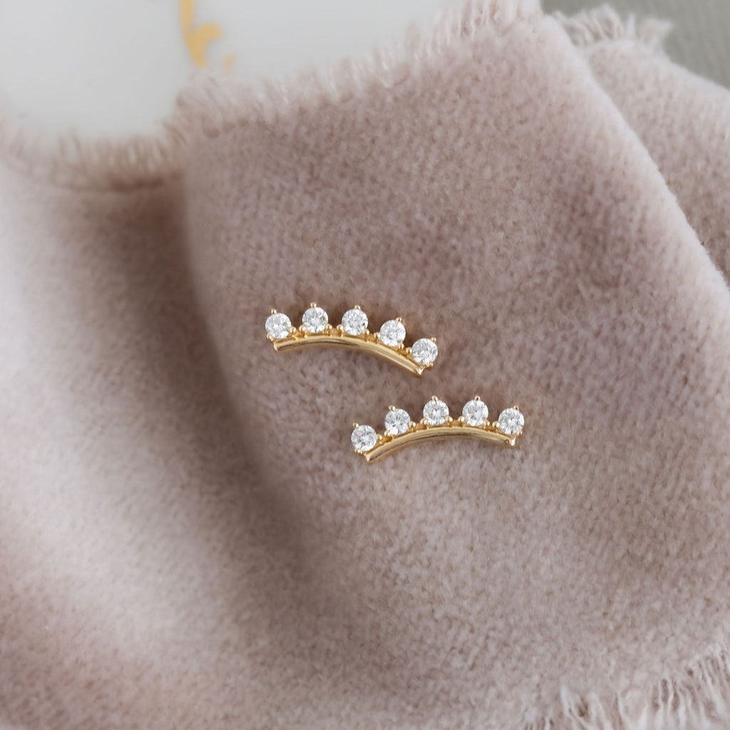 A pair of 14k gold stud earrings with round diamonds set on a curved bar, displayed on a grey fringed cloth.