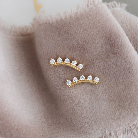 A pair of 14k gold stud earrings with round diamonds set on a curved bar, displayed on a grey fringed cloth.