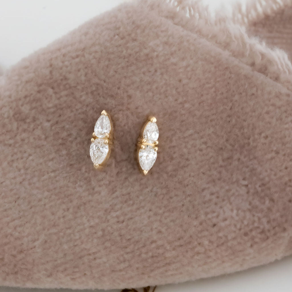 A pair of 14k yellow gold bar stud earrings with pear cut diamonds set in prongs, displayed on a beige soft surface.