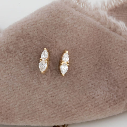 A pair of 14k yellow gold bar stud earrings with pear cut diamonds set in prongs, displayed on a beige soft surface.