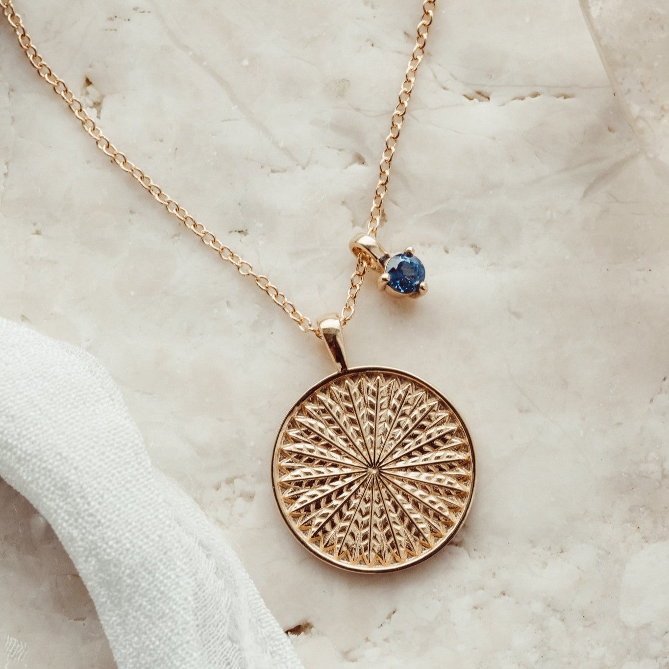A gold necklace with a textured circular pendant, displayed on a marble surface and paired with a gemstone charm