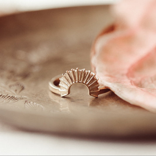 A 14k gold curved band ring with a design inspired by a dissent collar, displayed on a dish next to a pink fabric.