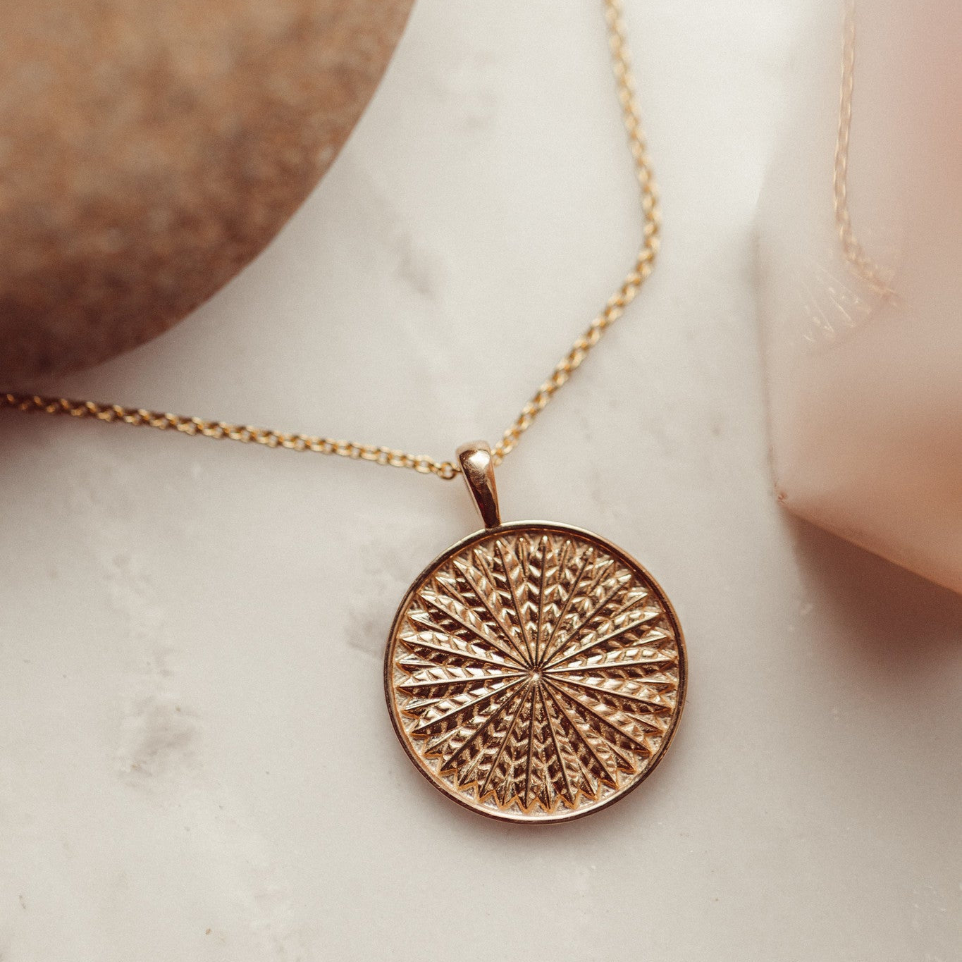 A gold necklace with a textured circular pendant, displayed on a marble surface.