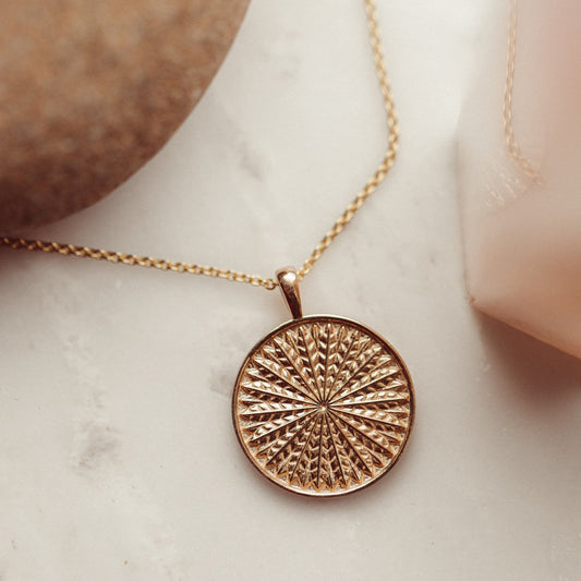 A gold necklace with a textured circular pendant, displayed on a marble surface.