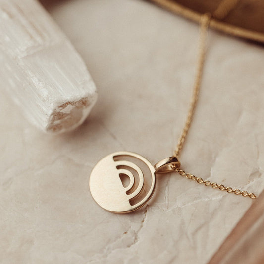 A 14k gold necklace with a circle pendant and rainbow cutout, displayed on an adjustable rolo chain, presented on a beige background.