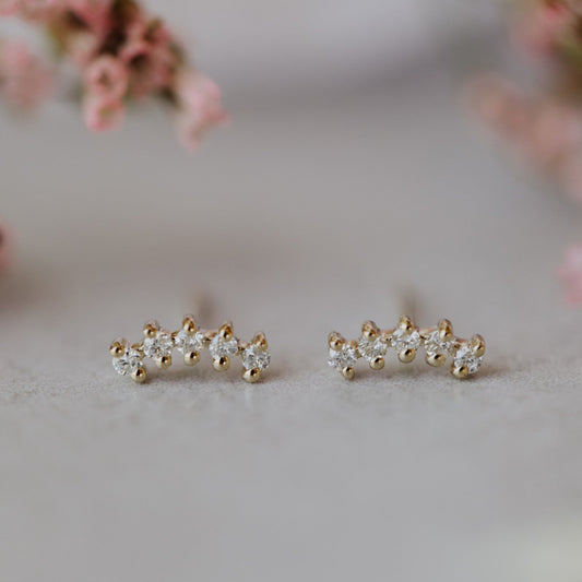 A pair of 14k yellow gold stud earrings featuring five round stones set in a curved starburst design, displayed against a floral background.