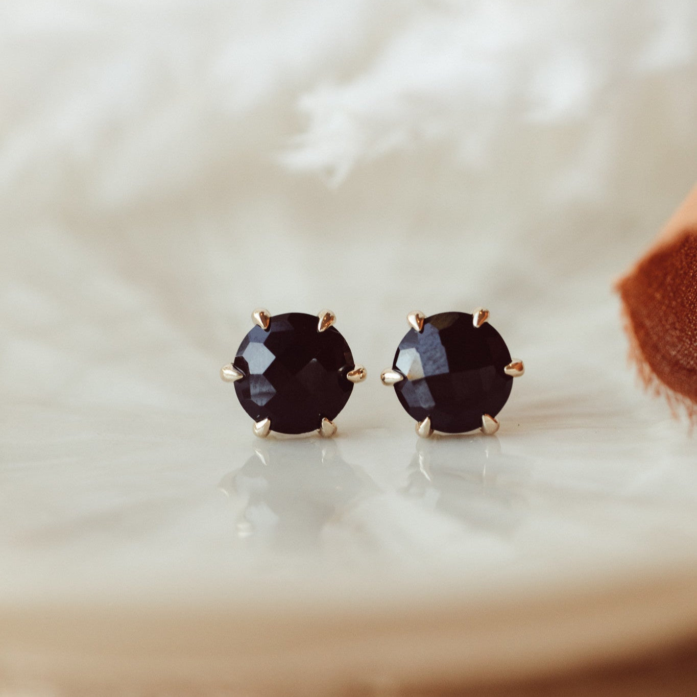 A pair of 14k gold stud earrings featuring round black spinel stones set in six prongs, displayed on a white platform with a textured background.