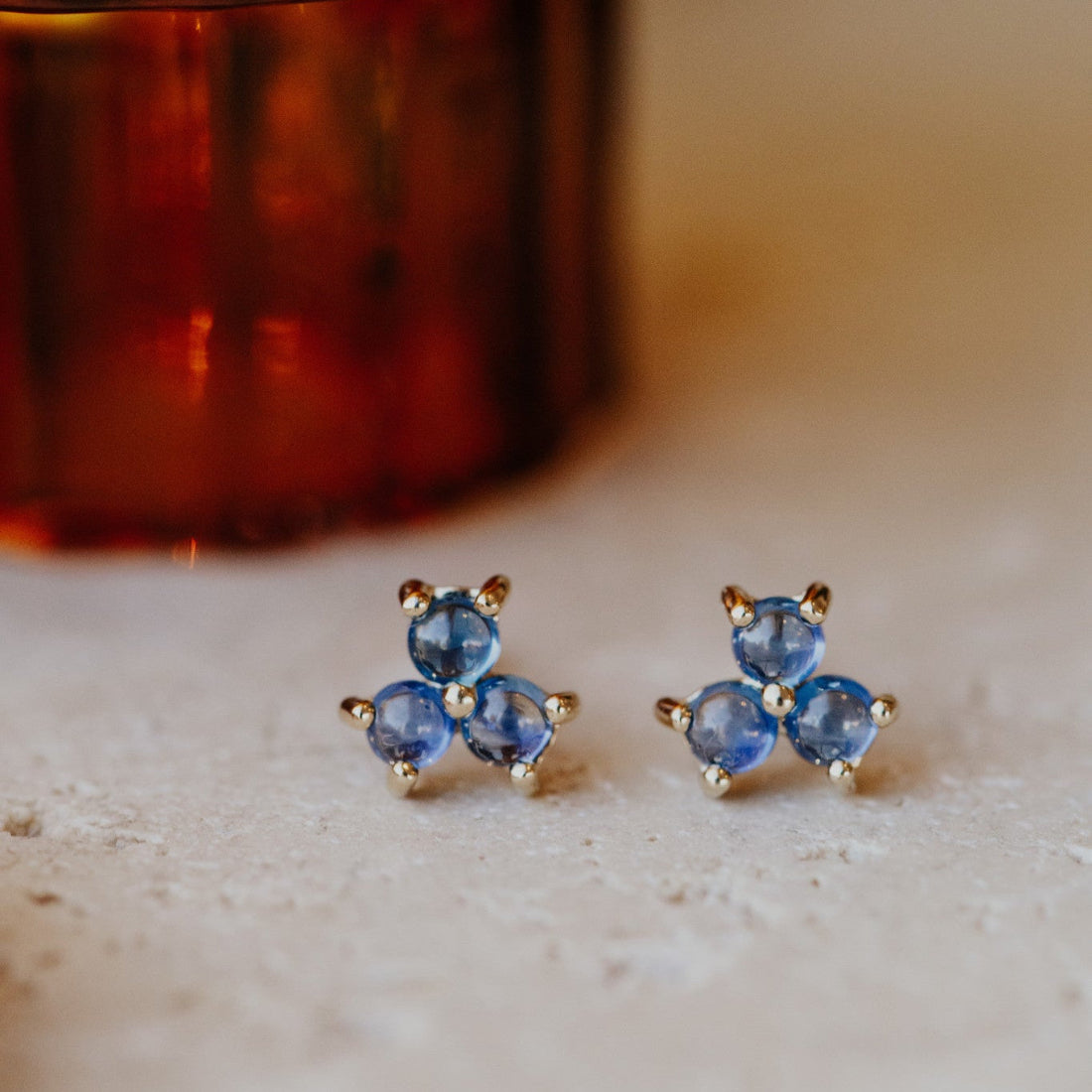 A pair of 14k yellow gold stud earrings featuring three round blue sapphires set in a cluster design, displayed against a reflective surface.