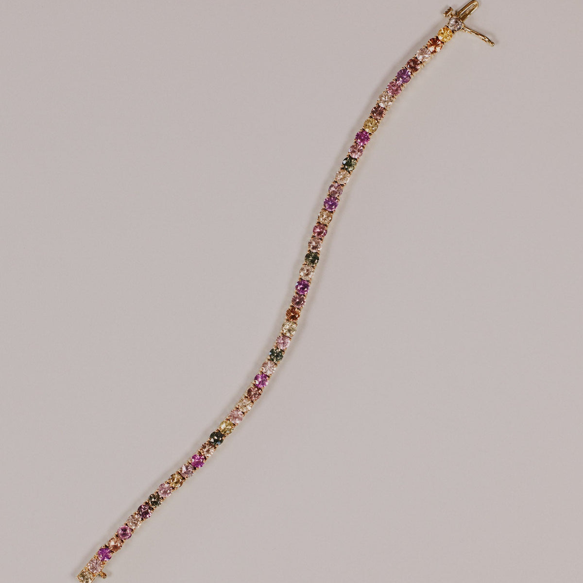 The 7 Inch 12.00 ctw Pastel Multicolored Natural Sapphire Tennis Bracelet in 14k yellow gold, featuring round cut natural sapphires in a mix of pastel pink, purple, green, orange, and champagne tones set in prongs, shown unclasped on a light gray surface.