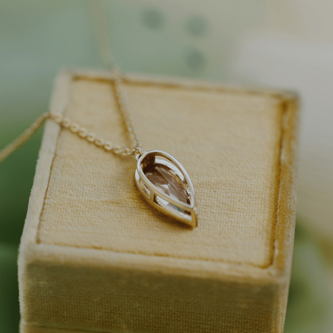 The back side of a 14k yellow gold pear-shaped morganite necklace in a bezel setting on a soft tan jewelry box with a textured background 