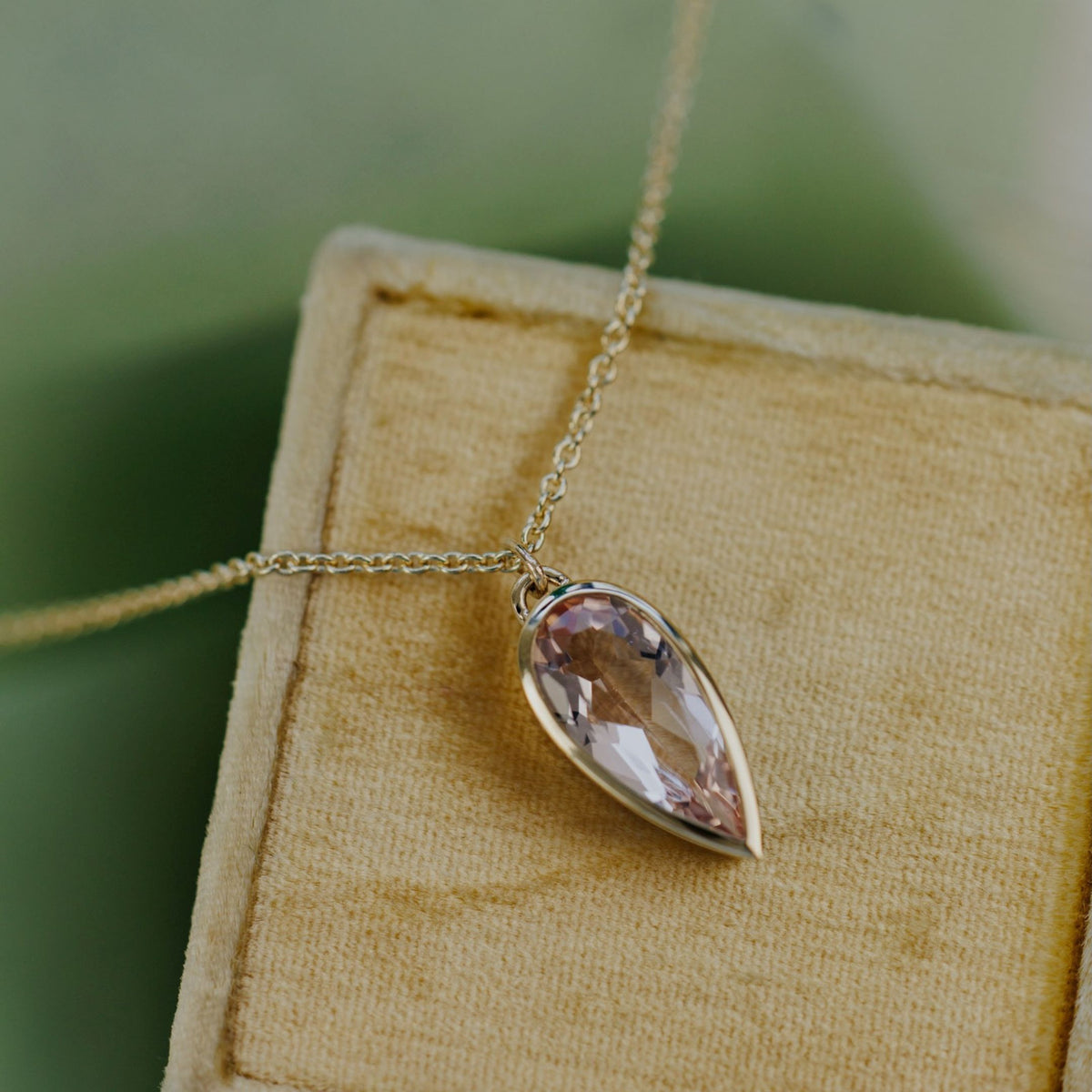 A 14k yellow gold pear-shaped morganite necklace in a bezel setting on a soft tan jewelry box with a textured background 