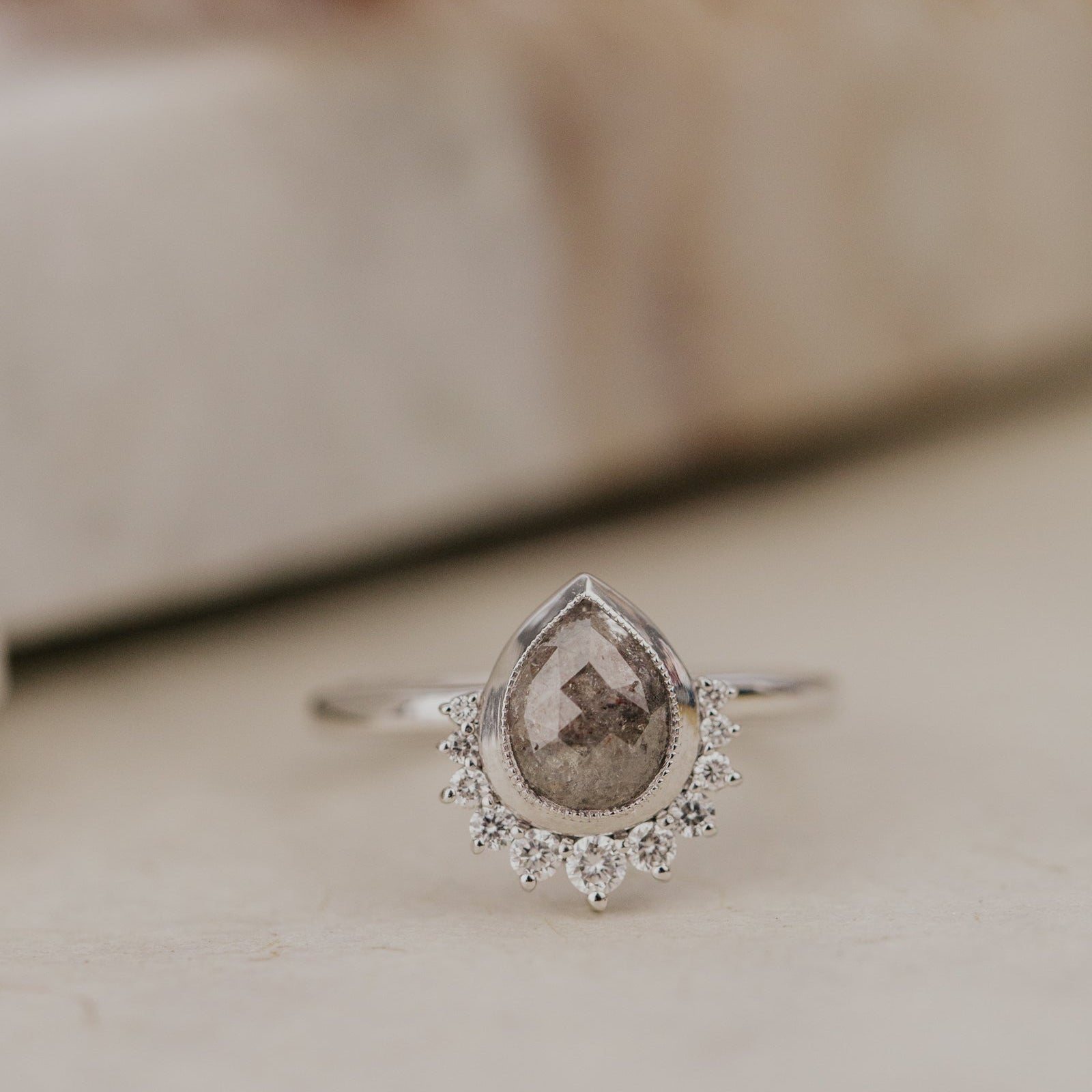 White Gold Acadia ring with a pear-shaped gray brown rustic diamond and diamond halo on a neutral background.