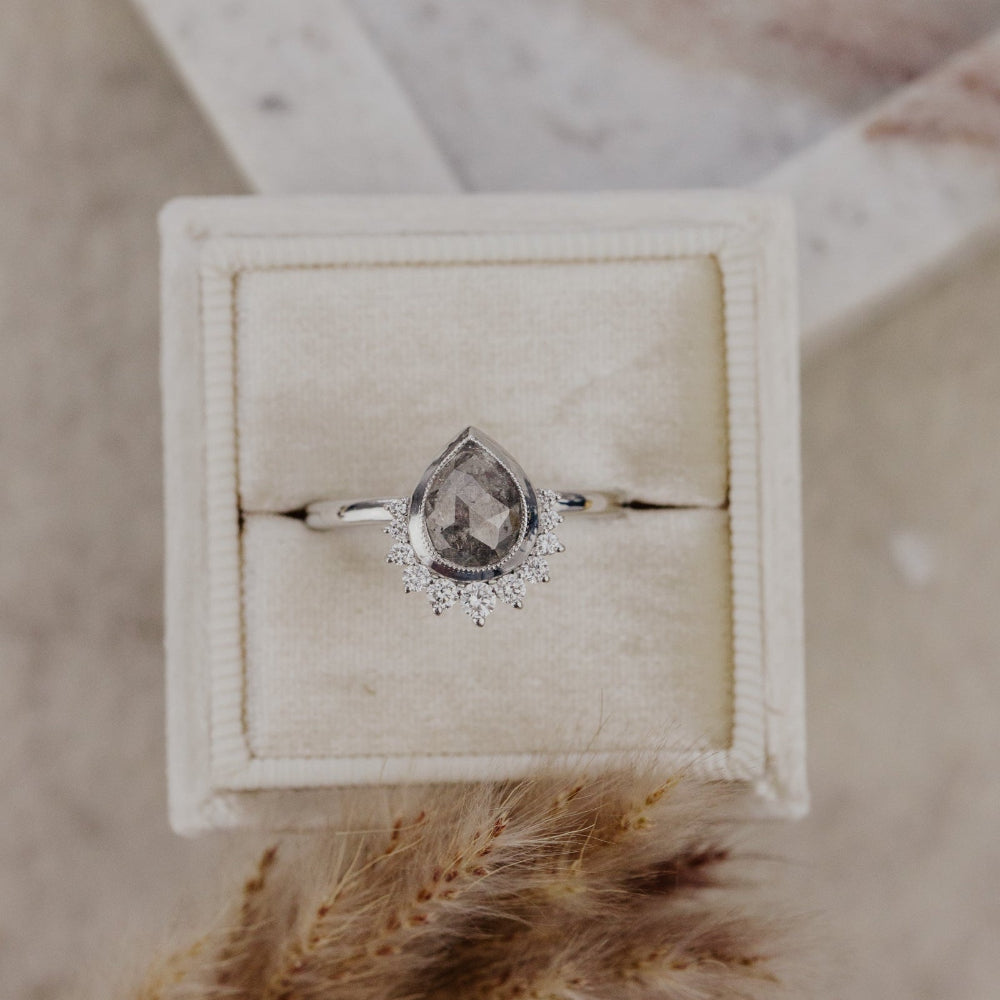 Pear-shaped rustic diamond ring in a decorative box on a textured surface with dried grass.