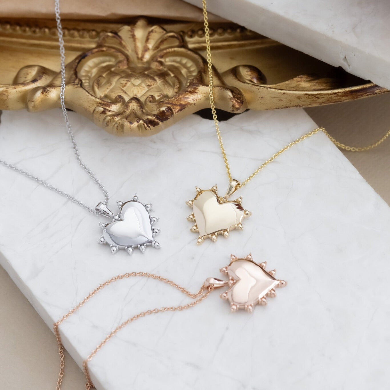 A collection of heart-shaped pendant necklaces in rose, yellow, and white gold, displayed on a white surface with an ornate golden frame in the background.