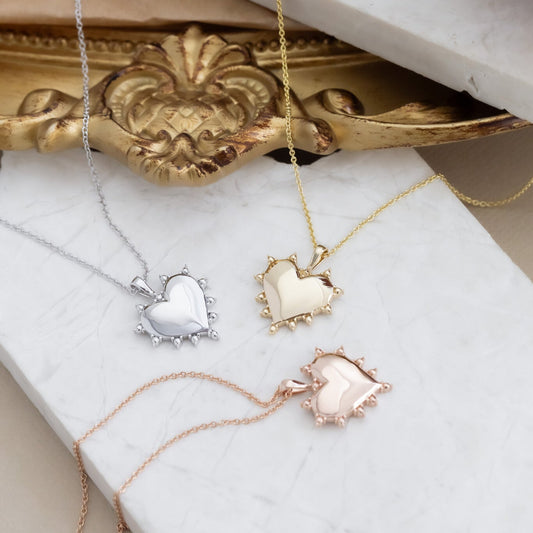 A collection of heart-shaped pendant necklaces in rose, yellow, and white gold, displayed on a white surface with an ornate golden frame in the background.