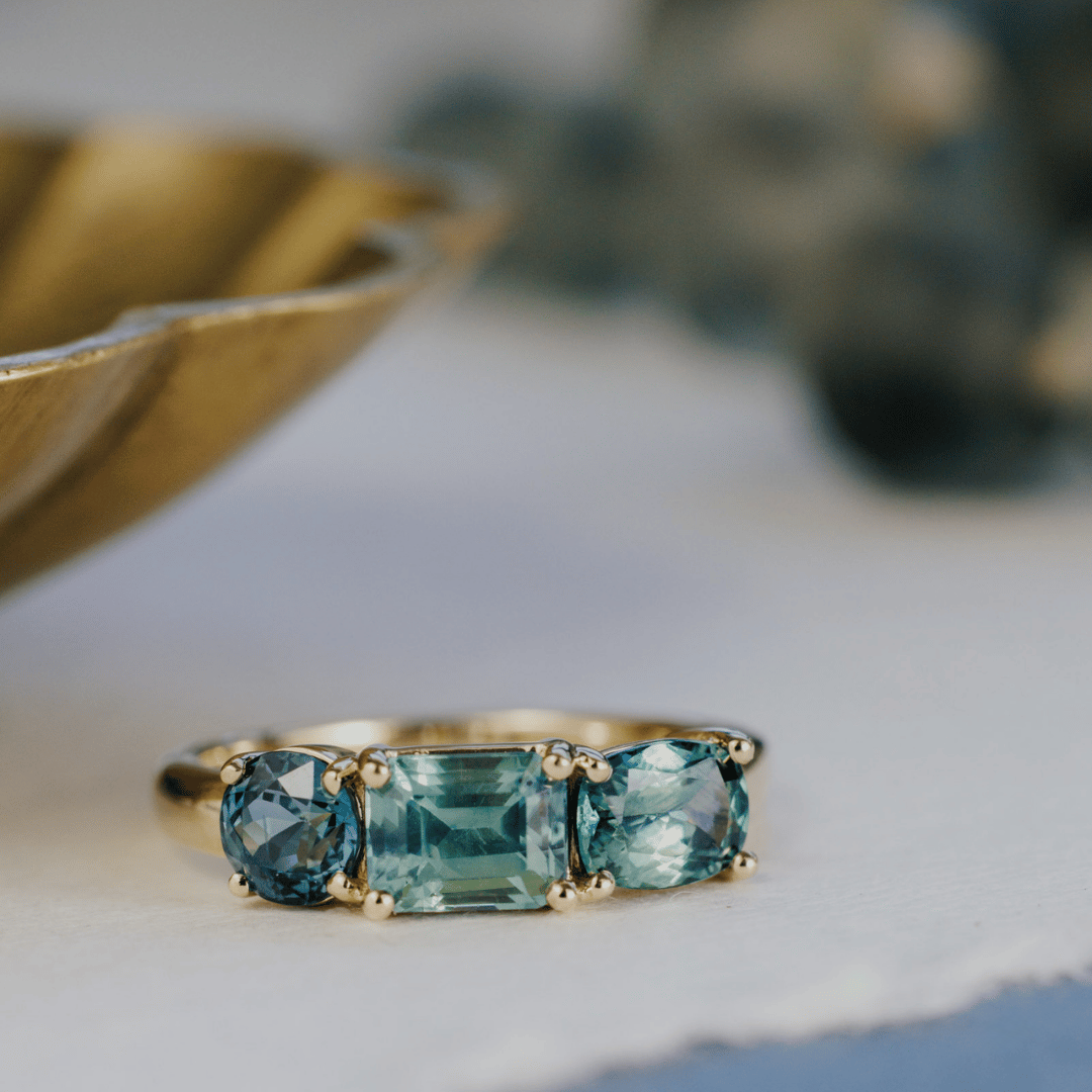 A 14k gold ring featuring three blue sapphire gemstones, displayed on a textured white surface with a soft-focus background.
