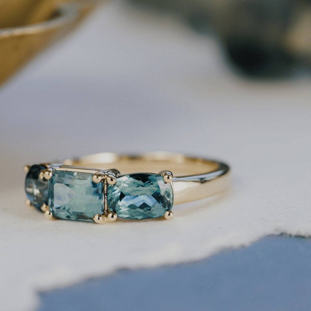 A 14k gold ring featuring three blue sapphire gemstones, displayed on a textured white surface with a soft-focus background. Ring is shifted to the side, featuring one of the side stones as the main focus. 