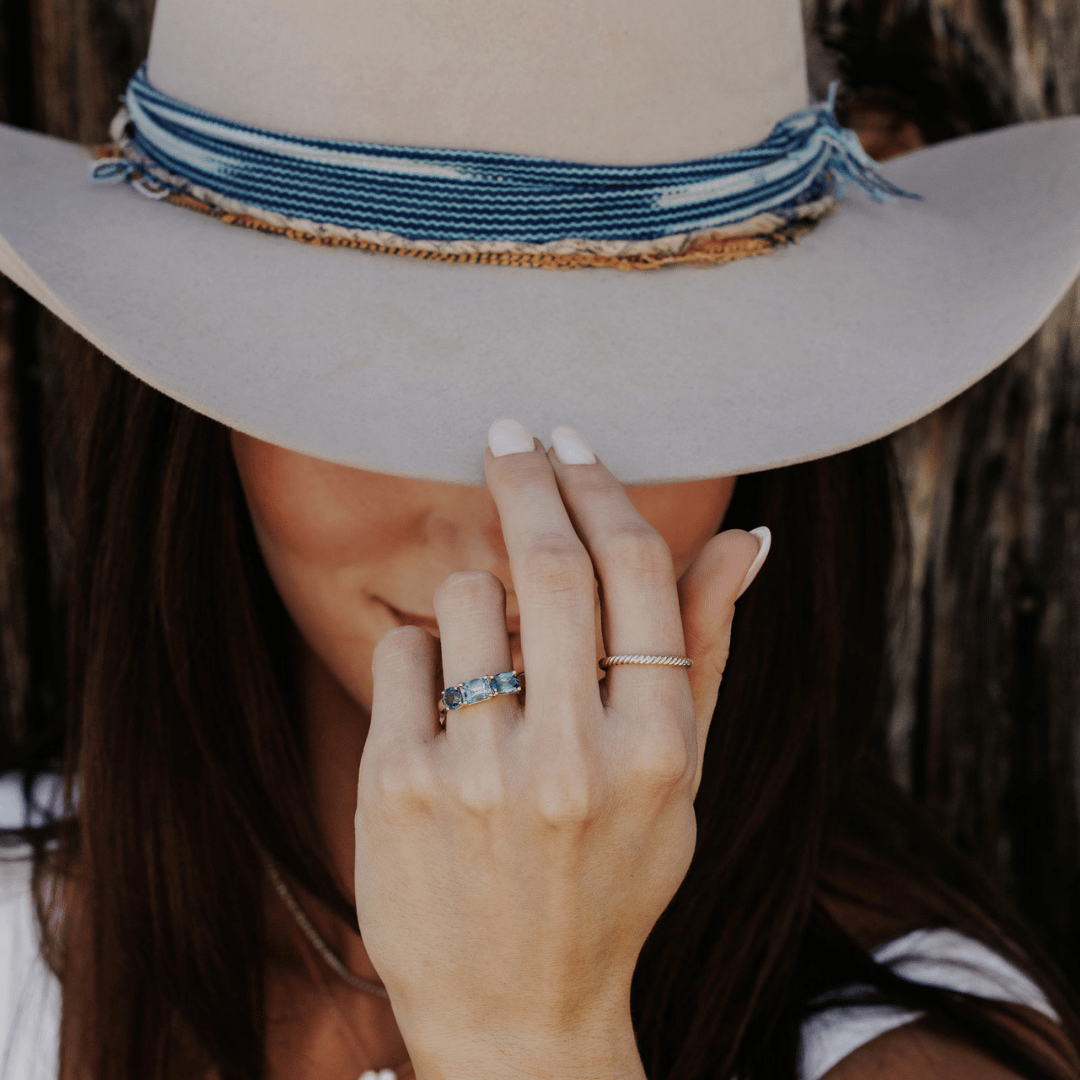 A 14k gold ring featuring three blue sapphire gemstones, displayed on a the ring finger of a hand model tipping her hat. 