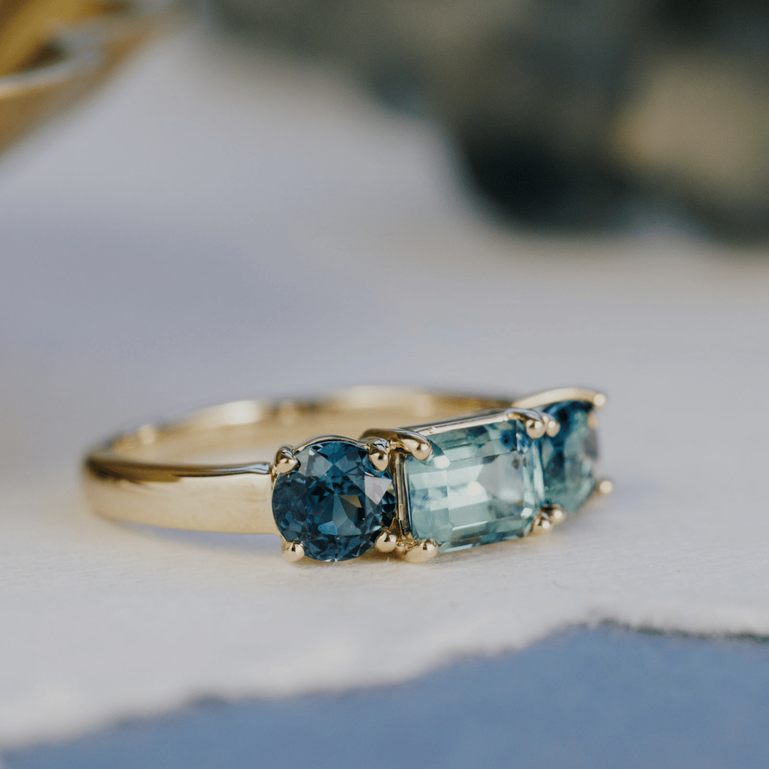 A 14k gold ring featuring three blue sapphire gemstones, displayed on a textured white and blue surface with a soft-focus background. Ring is shifted slightly to feature the side stones.