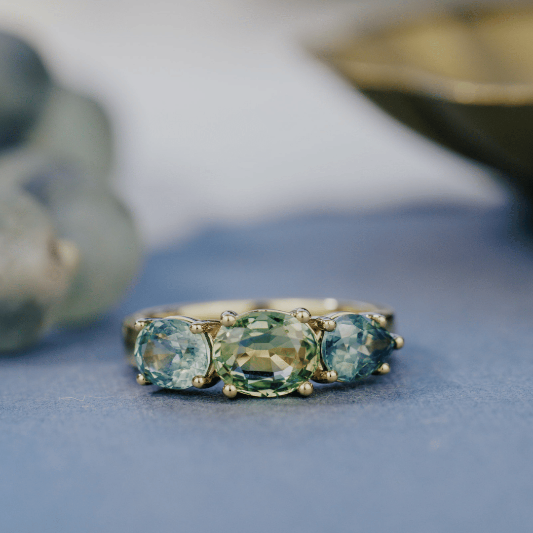 A 14k gold ring featuring three blue sapphire gemstones, displayed on a textured blue surface with a soft-focus background.