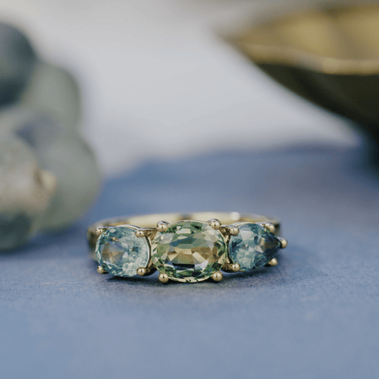 A 14k gold ring featuring three blue sapphire gemstones, displayed on a textured blue surface with a soft-focus background.