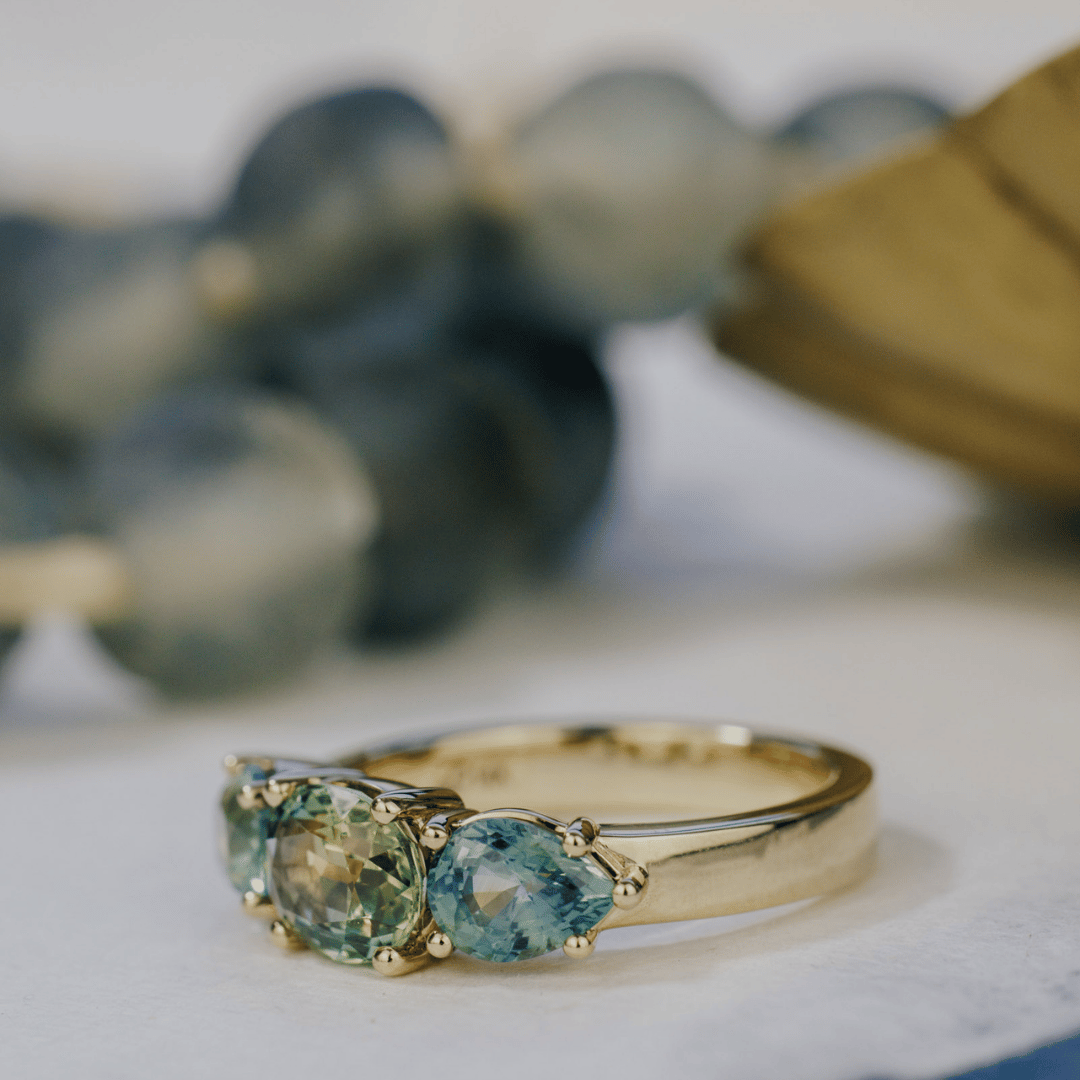 A 14k gold ring featuring three blue sapphire gemstones, displayed on a textured white surface with a soft-focus background. Ring is set to the side with the pear shaped sapphire mostly featured. 
