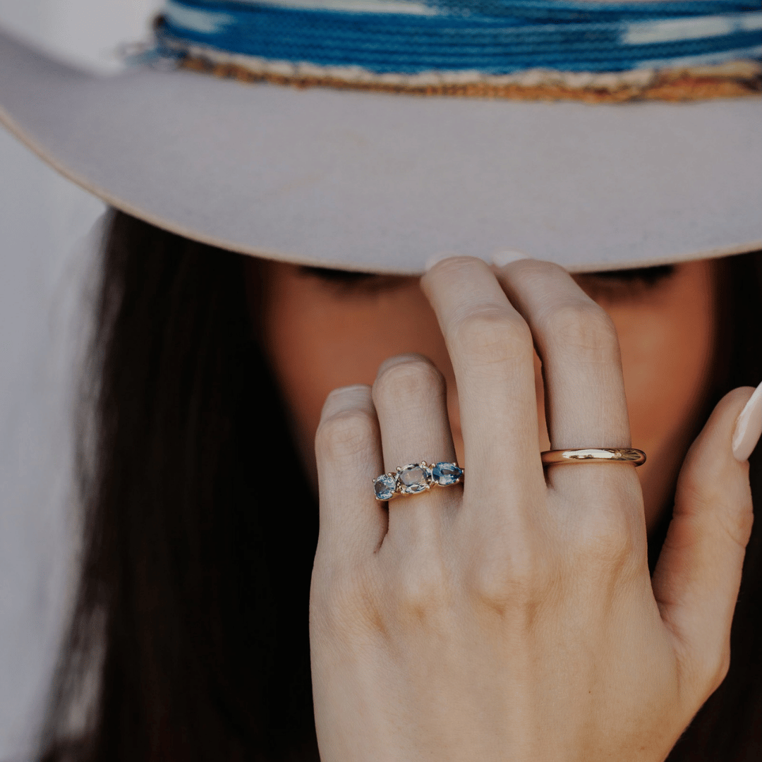 A 14k gold ring featuring three blue sapphire gemstones, displayed on the ring finger of a hand model who is tipping the brim of her hat. 
