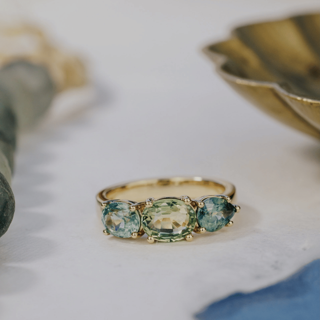 A 14k gold ring featuring three blue sapphire gemstones, displayed on a textured white surface with a soft-focus background.