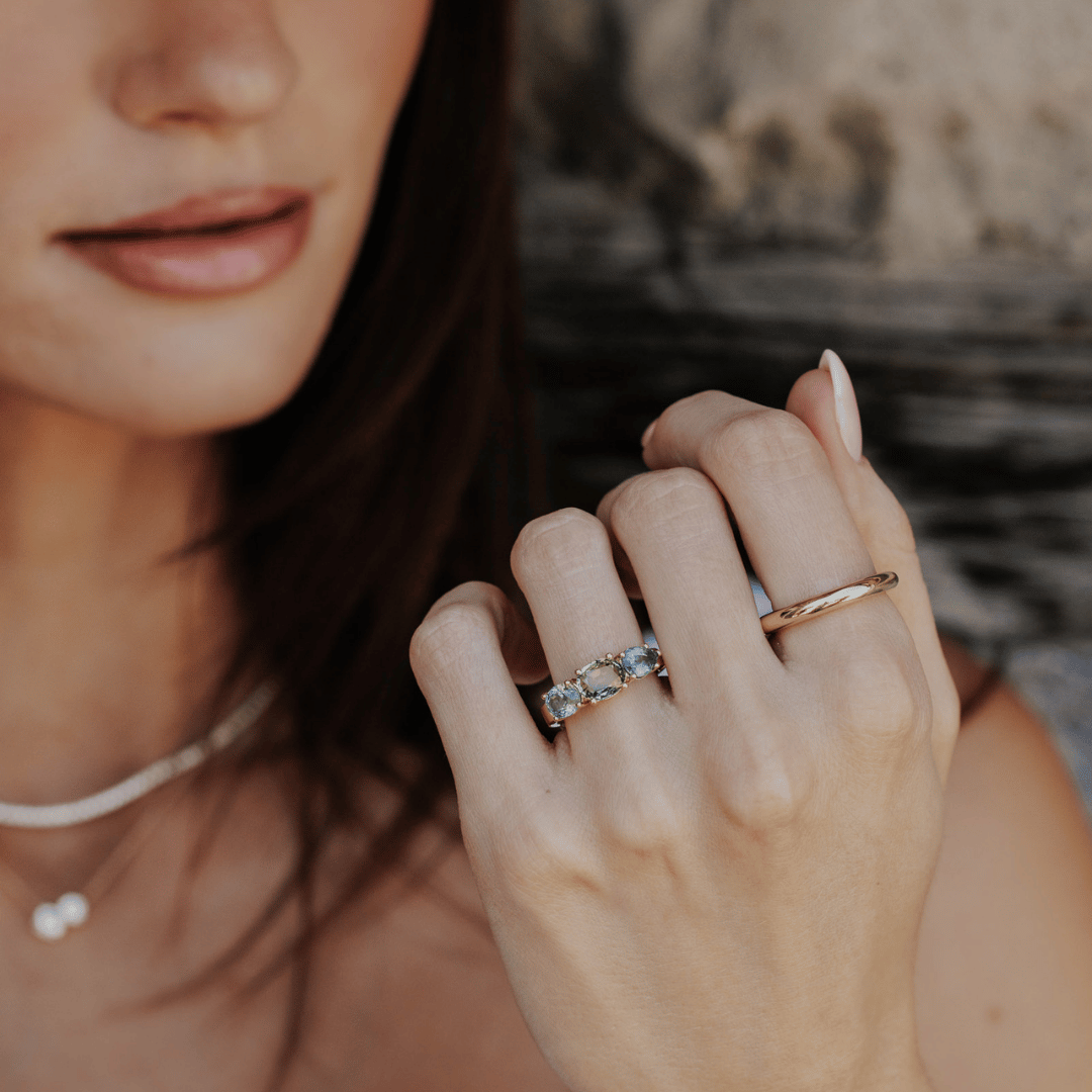 A 14k gold ring featuring three blue sapphire gemstones, displayed on the ring finger of a hand model with a soft-focus background.
