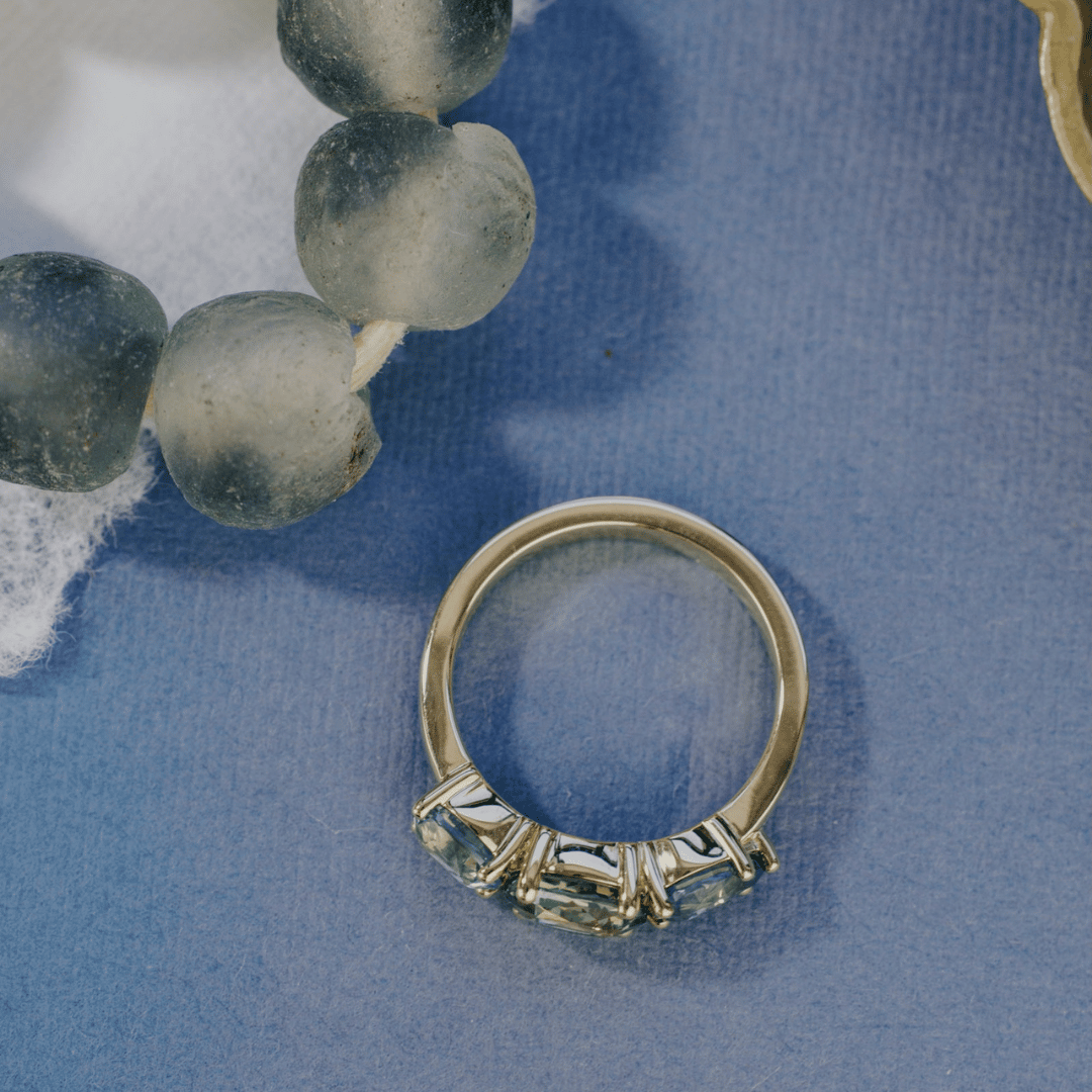 An overhead view of a 14k gold ring featuring three blue sapphire gemstones, displayed on a textured blue surface, sat next to grey beads with a soft-focus background.