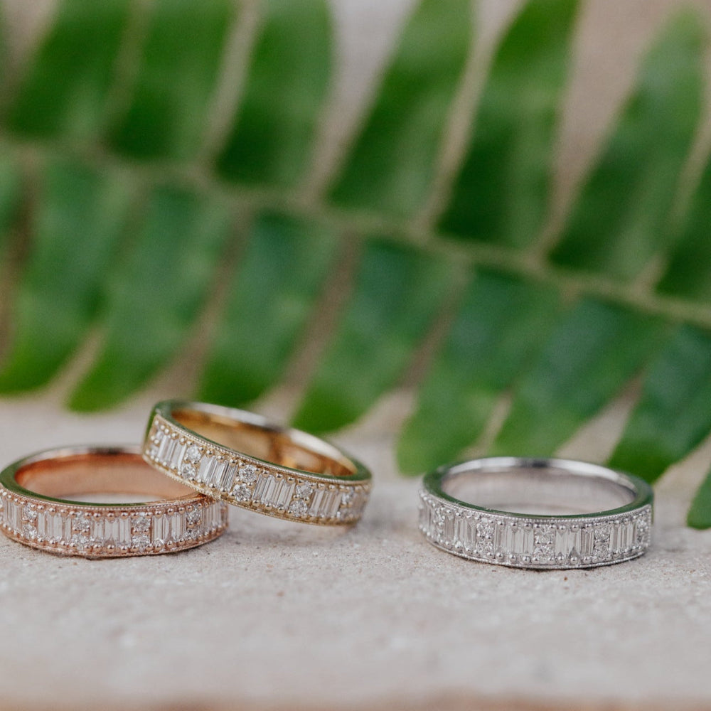 Three diamond bands in 14k yellow, white, and rose gold on a stone surface with green foliage in the background