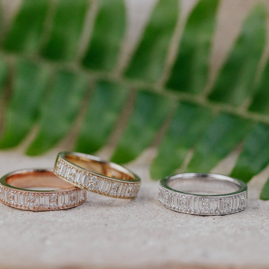 Three diamond bands in 14k yellow, white, and rose gold on a stone surface with green foliage in the background