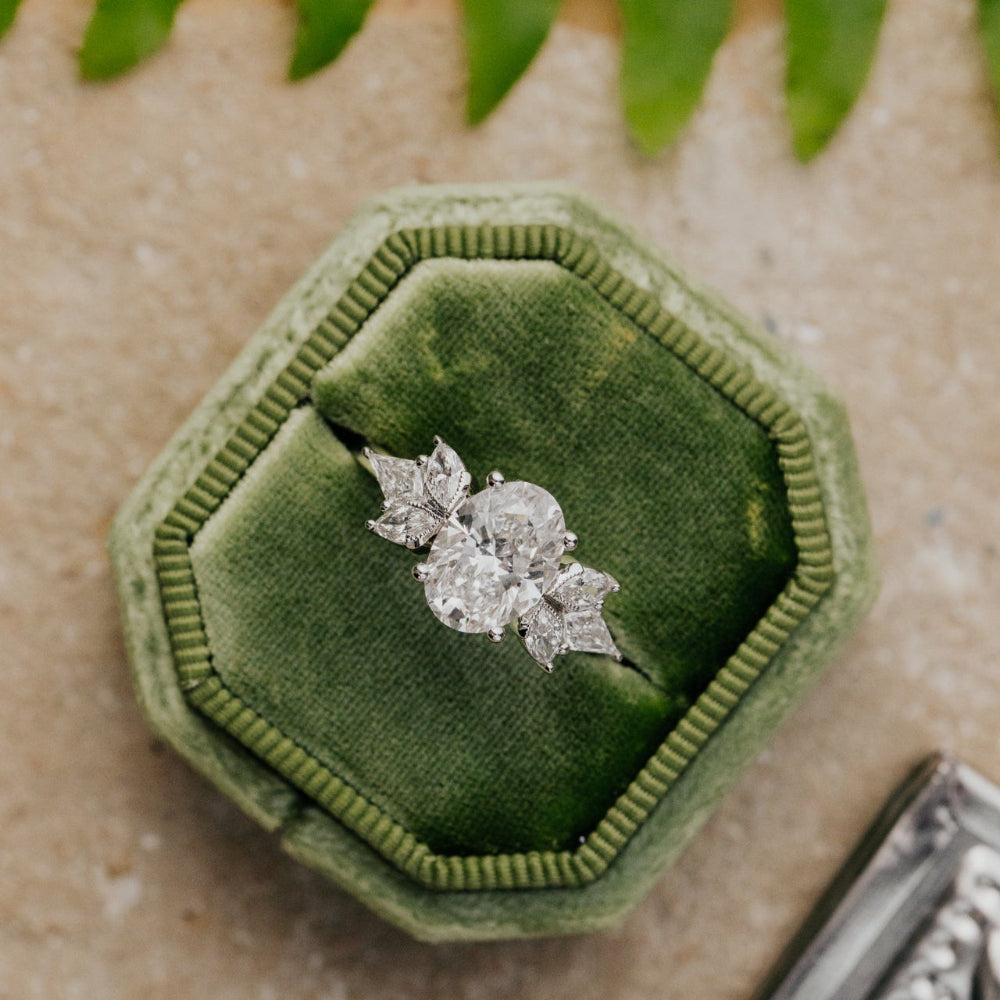 Oval diamond ring in a green velvet box on a textured surface with leaves.