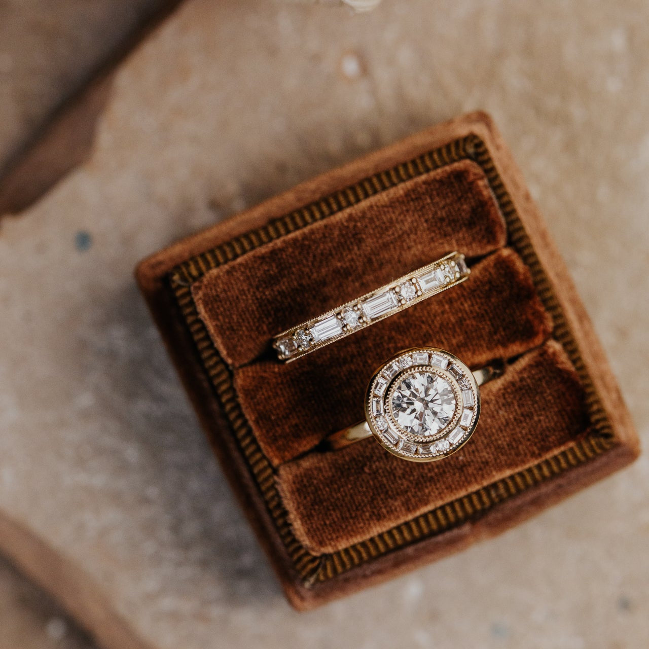 Two diamond rings in a brown velvet box on a textured surface.