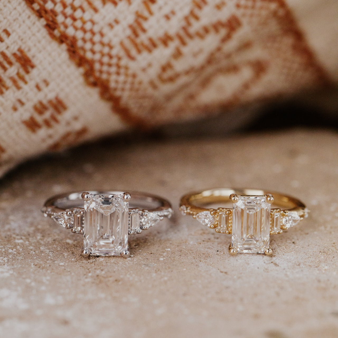 Two diamond emerald cut rings, one white gold and one in yellow gold, on a textured surface.