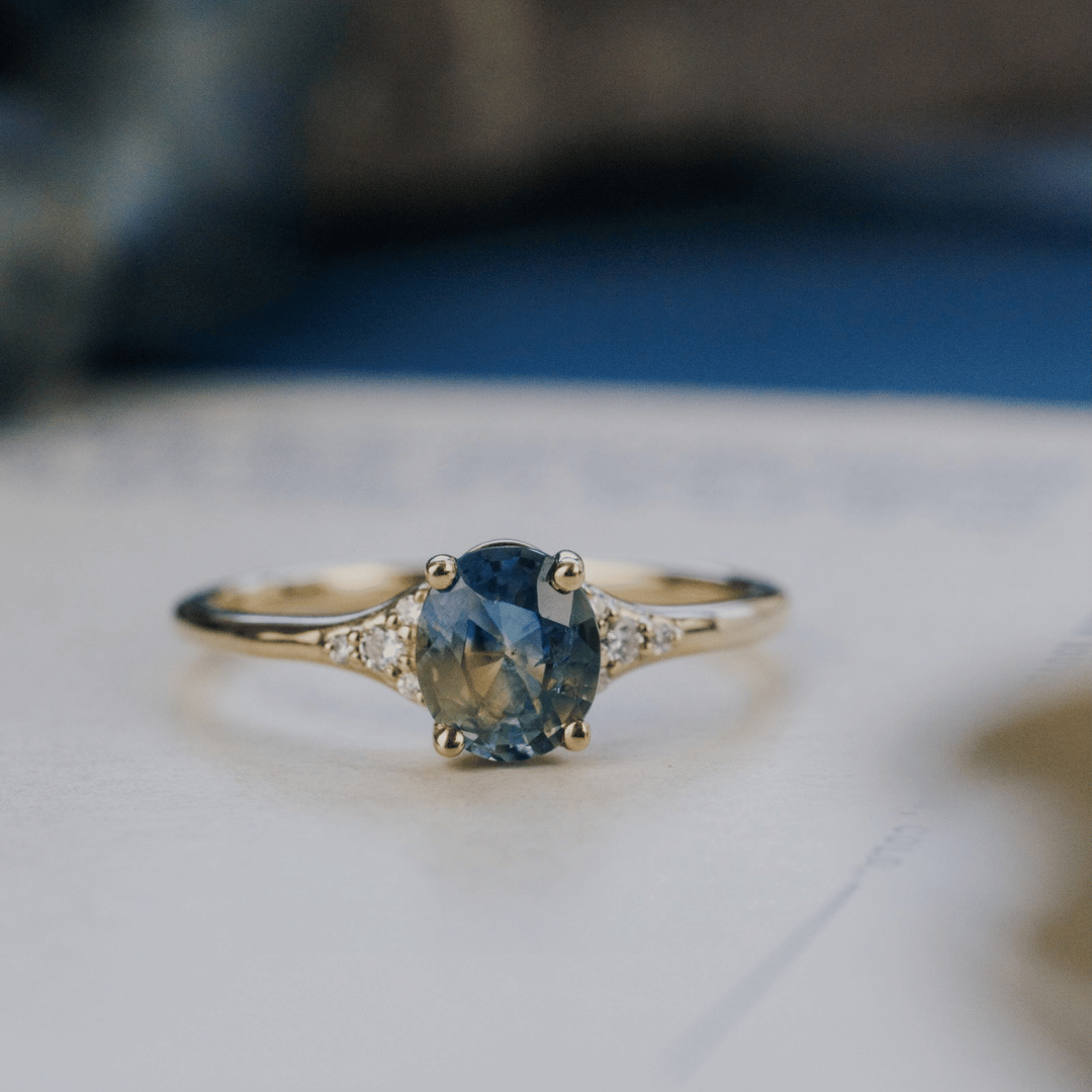 A 14k gold ring featuring a blue sapphire gemstone viewed from straight on, displayed on a textured white surface with a soft-focus background.