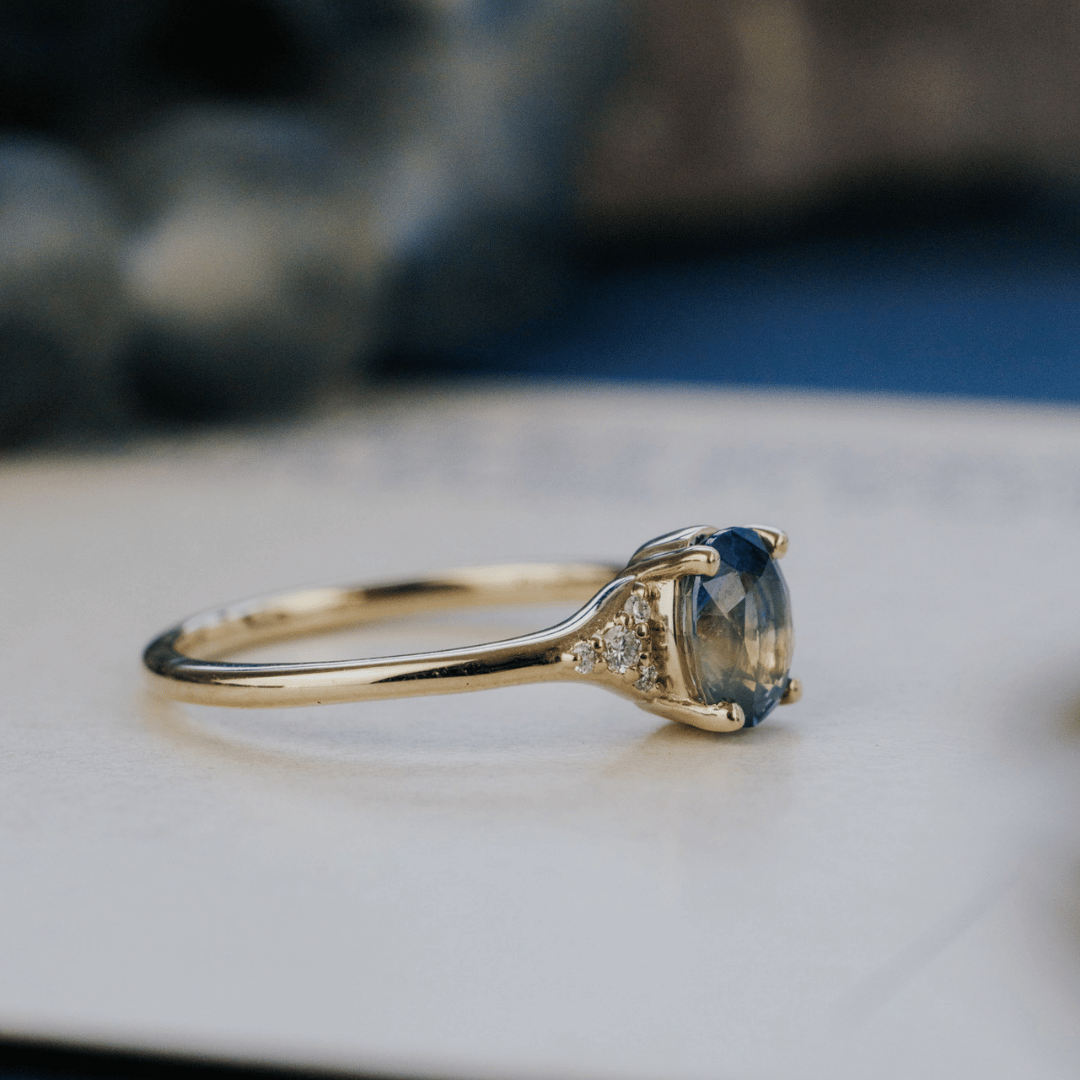 A 14k gold ring featuring a blue sapphire gemstone viewed from the side, displayed on a textured white surface with a soft-focus background.