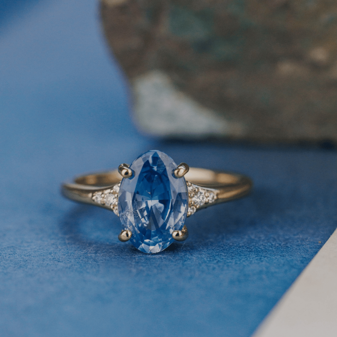 A 14k gold ring featuring a blue sapphire gemstone viewed from straight on, displayed on a textured blue surface with a soft-focus background.