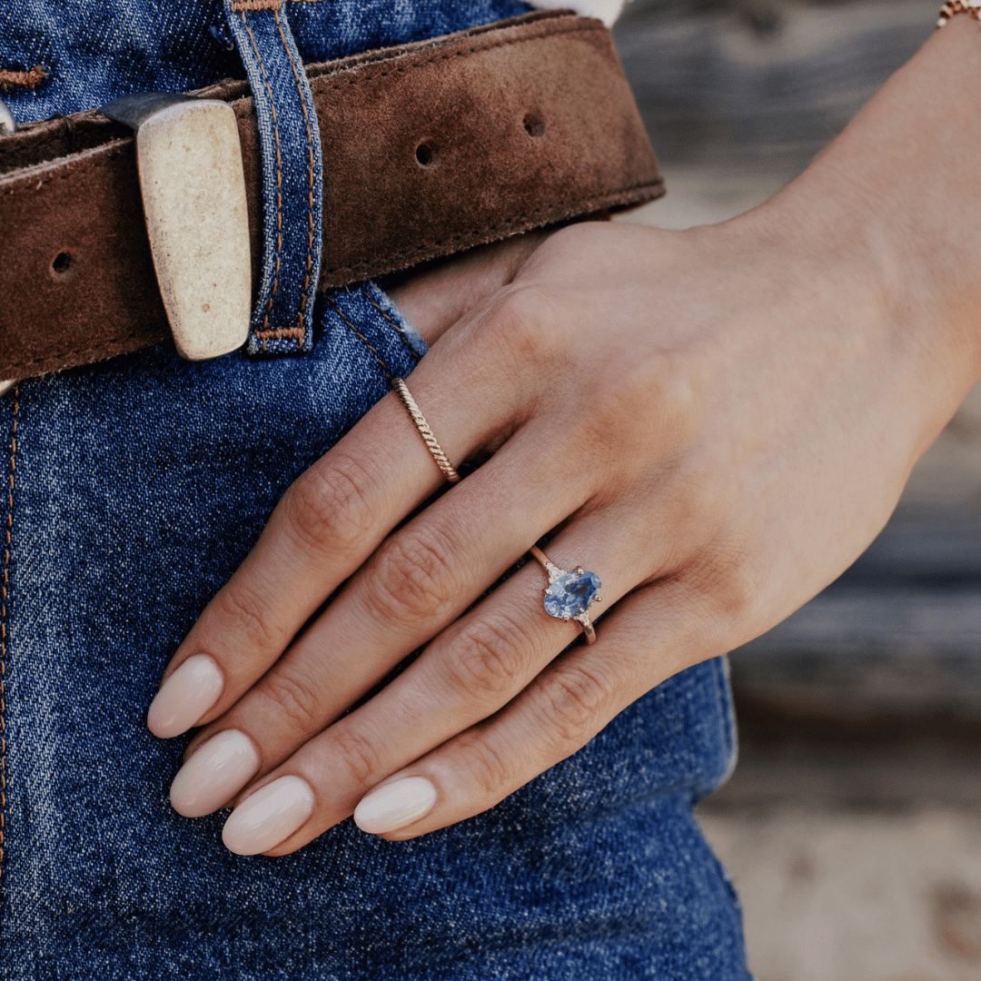 A 14k gold ring featuring a blue sapphire gemstone viewed the ring finger of a hand model. 