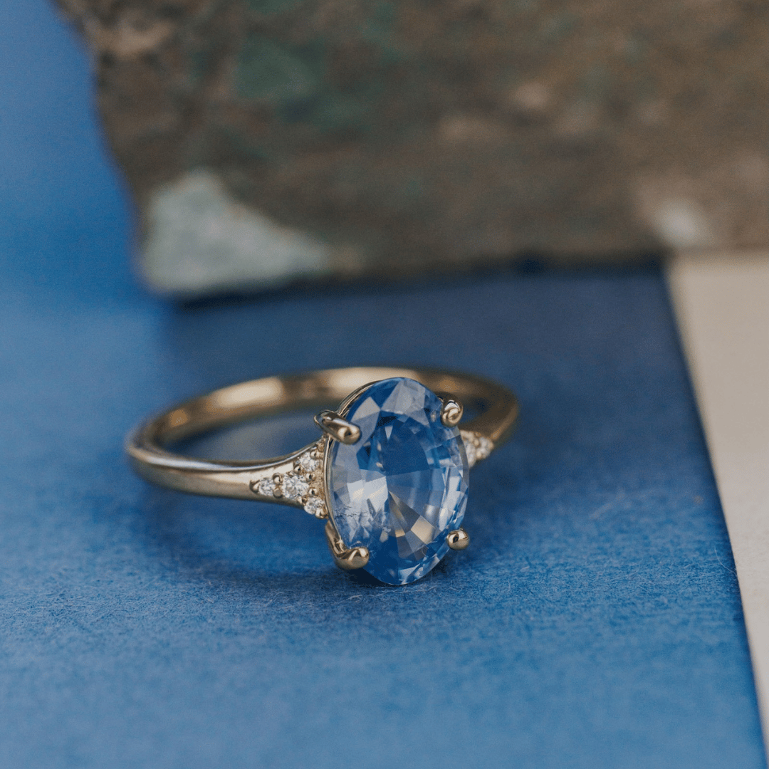 A 14k gold ring featuring a blue sapphire gemstone, displayed on a textured blue surface with a soft-focus background.