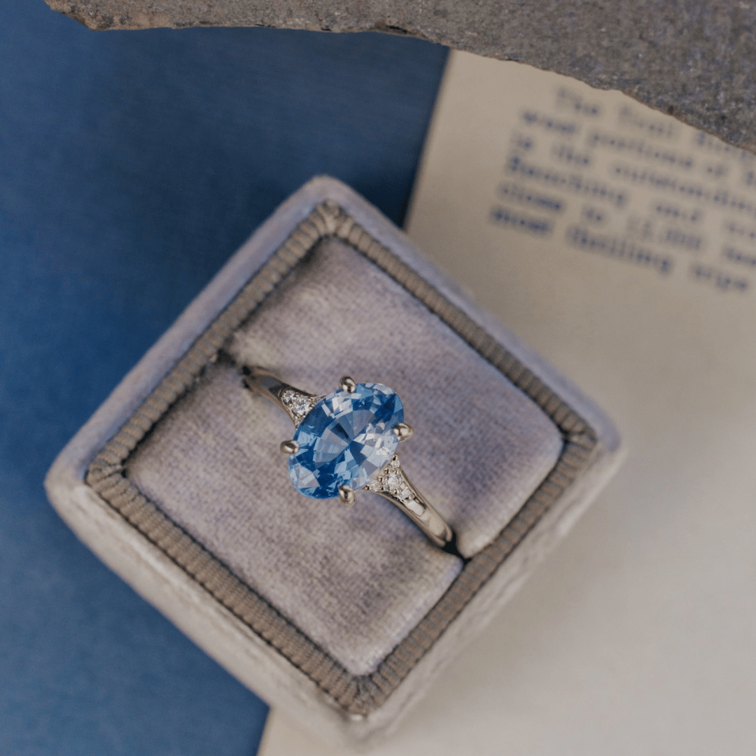 A 14k gold ring featuring a blue sapphire gemstone viewed from a. ring box, displayed on a textured white and blue surface with a soft-focus background.