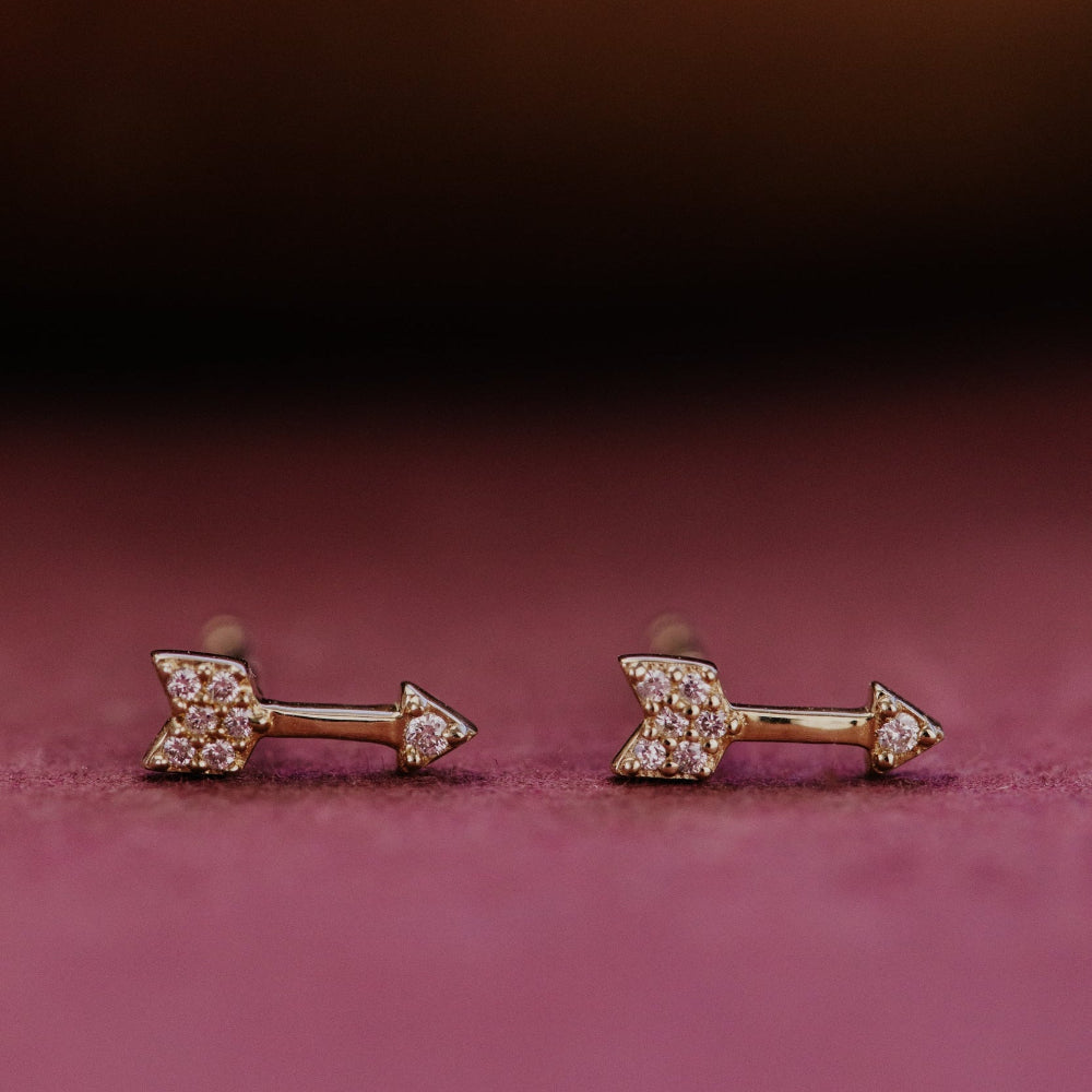 Pair of gold arrow-shaped diamond stud earrings with gemstones on a pink background.