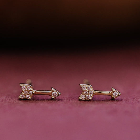 Pair of gold arrow-shaped diamond stud earrings with gemstones on a pink background.