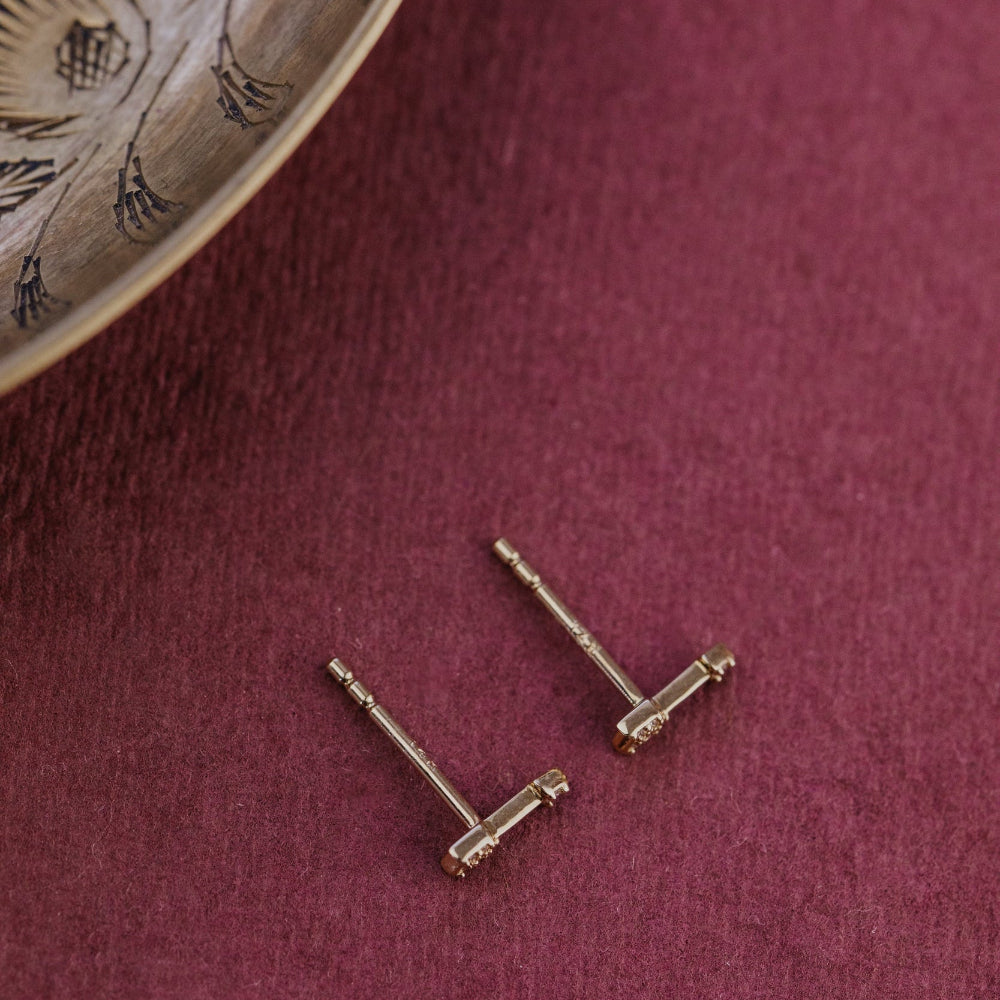 Gold stud earrings on a maroon surface with a wooden coaster in the corner.