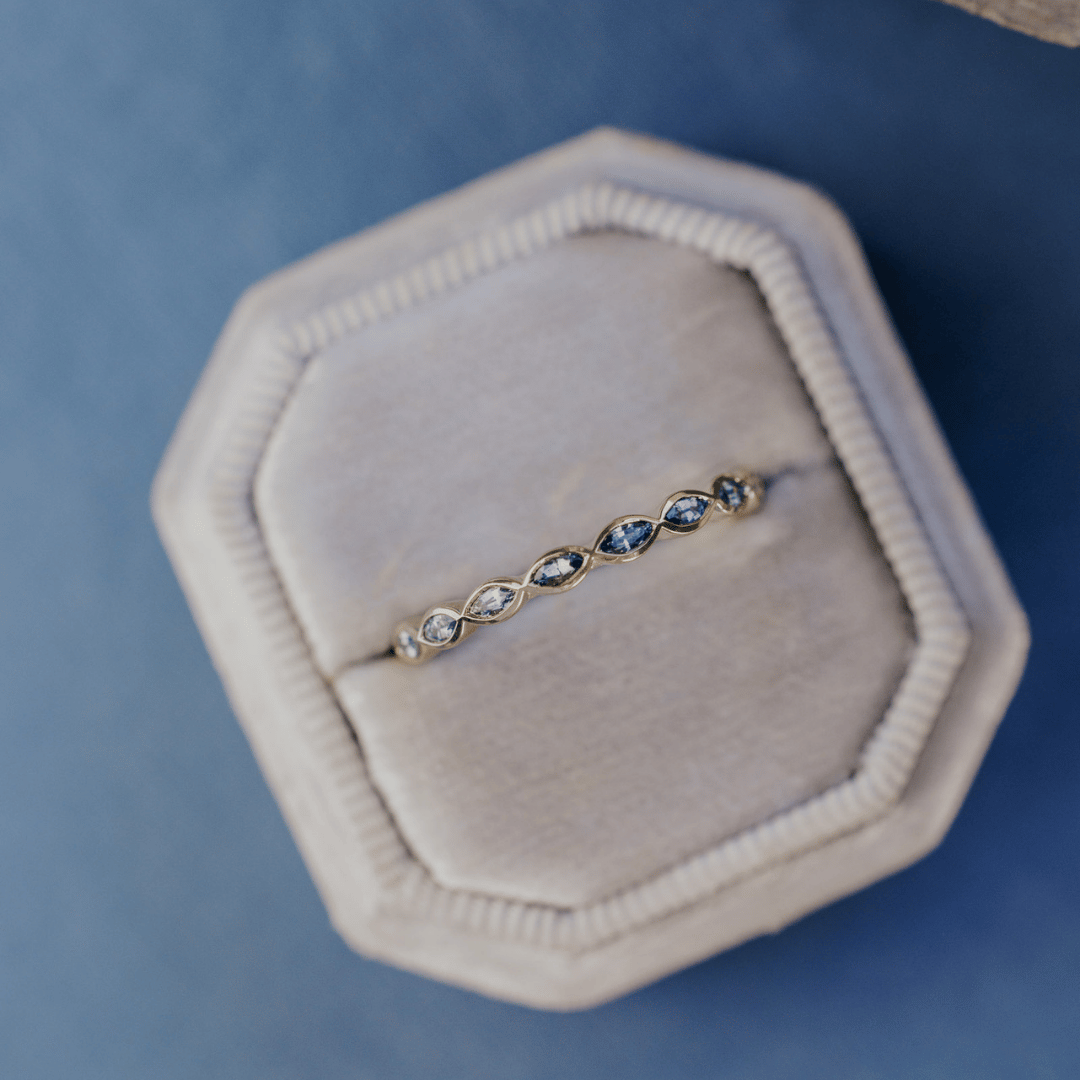 A 14k gold ring featuring blue marquise sapphire gemstones displayed in a velvet ring box on a blue surface with a soft-focus background.