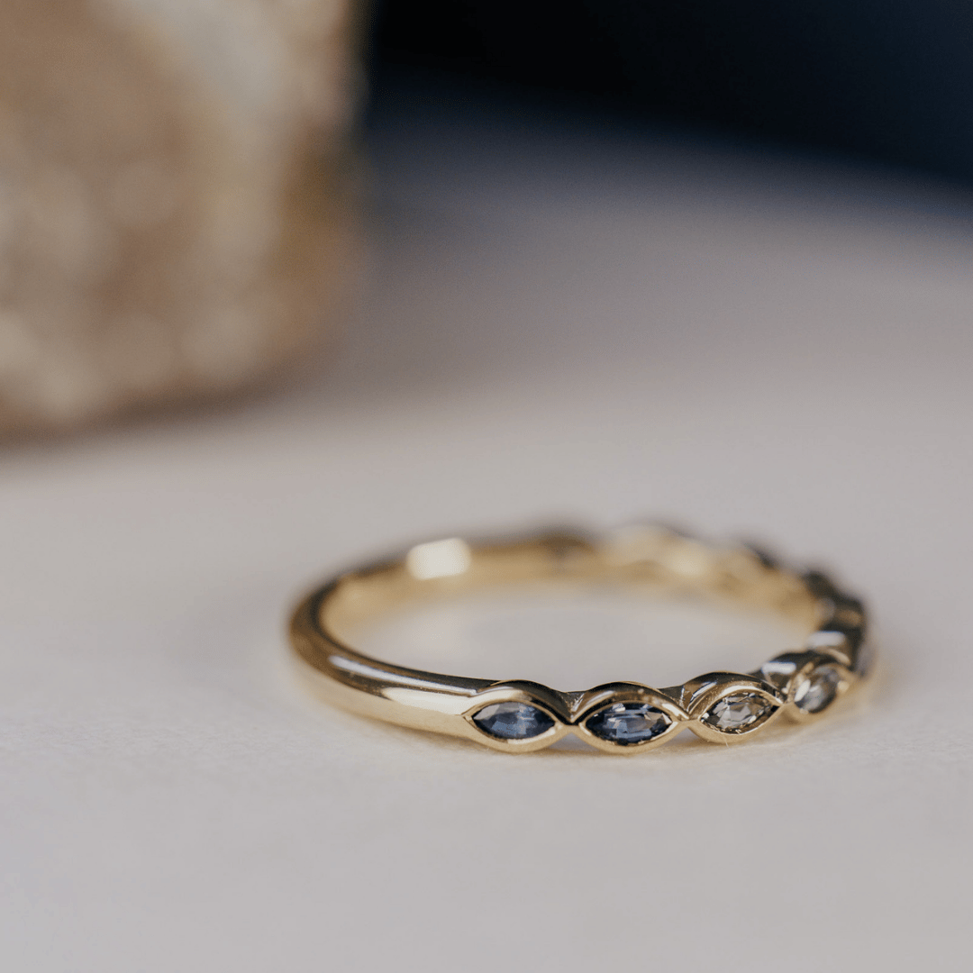 A 14k gold ring featuring blue marquise sapphire gemstones from an angled POV displayed on a white surface with a soft-focus background.